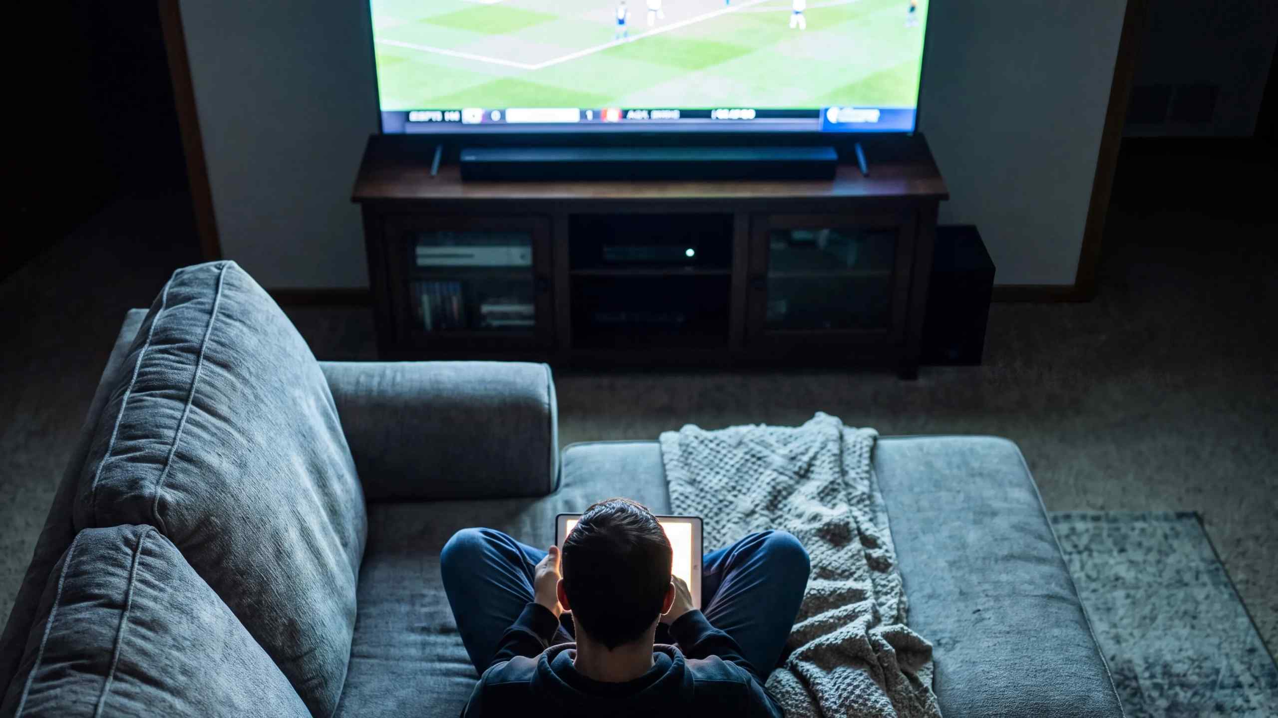 A man watching soccer on TV while using his laptop on the couch.